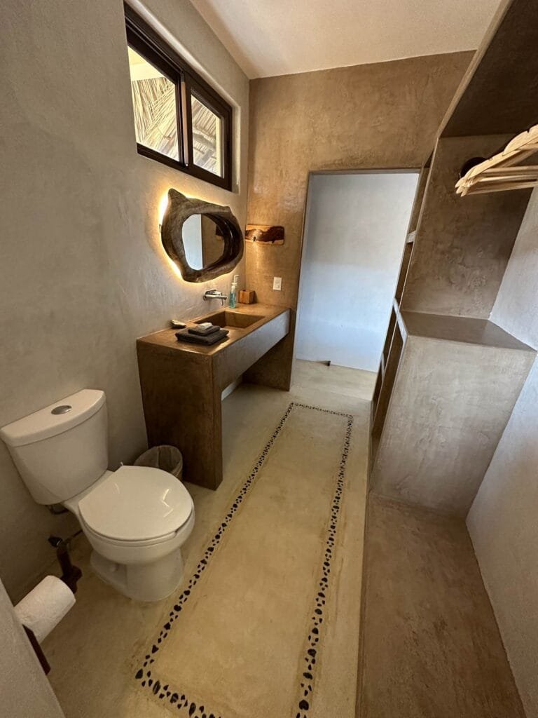 Baño privado de la Suite 1 con tocador de concreto, espejo iluminado de madera orgánica, inodoro y vestidor abierto