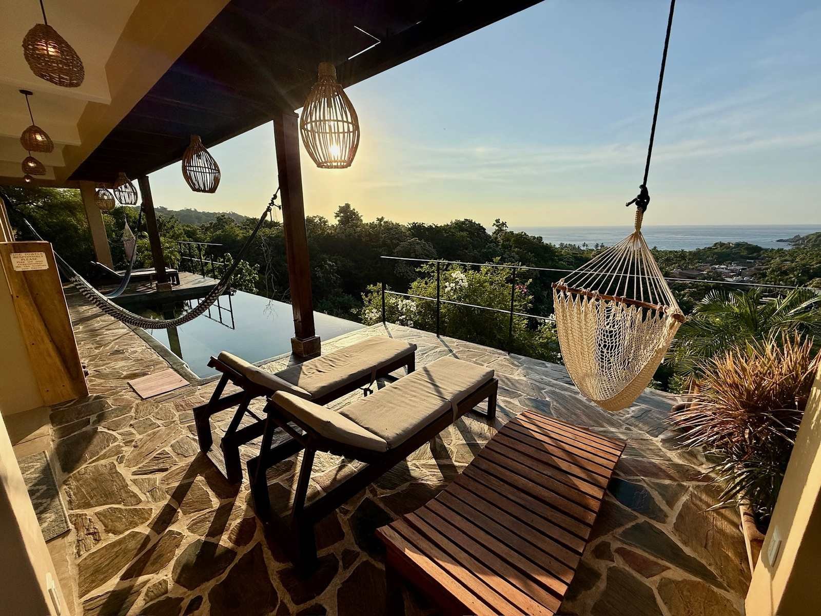 Terraza con camastros, silla colgante y alberca infinita con luz dorada en Casa La Vista, Mazunte