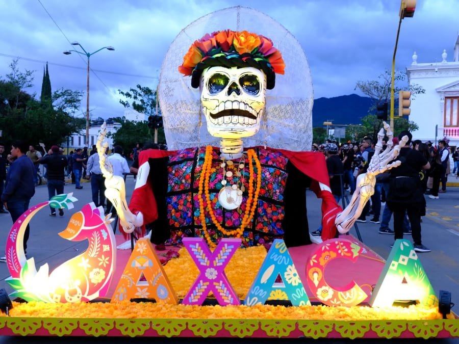 Carro alegórico con una catrina gigante y letras que dicen Oaxaca en un desfile de Día de Muertos