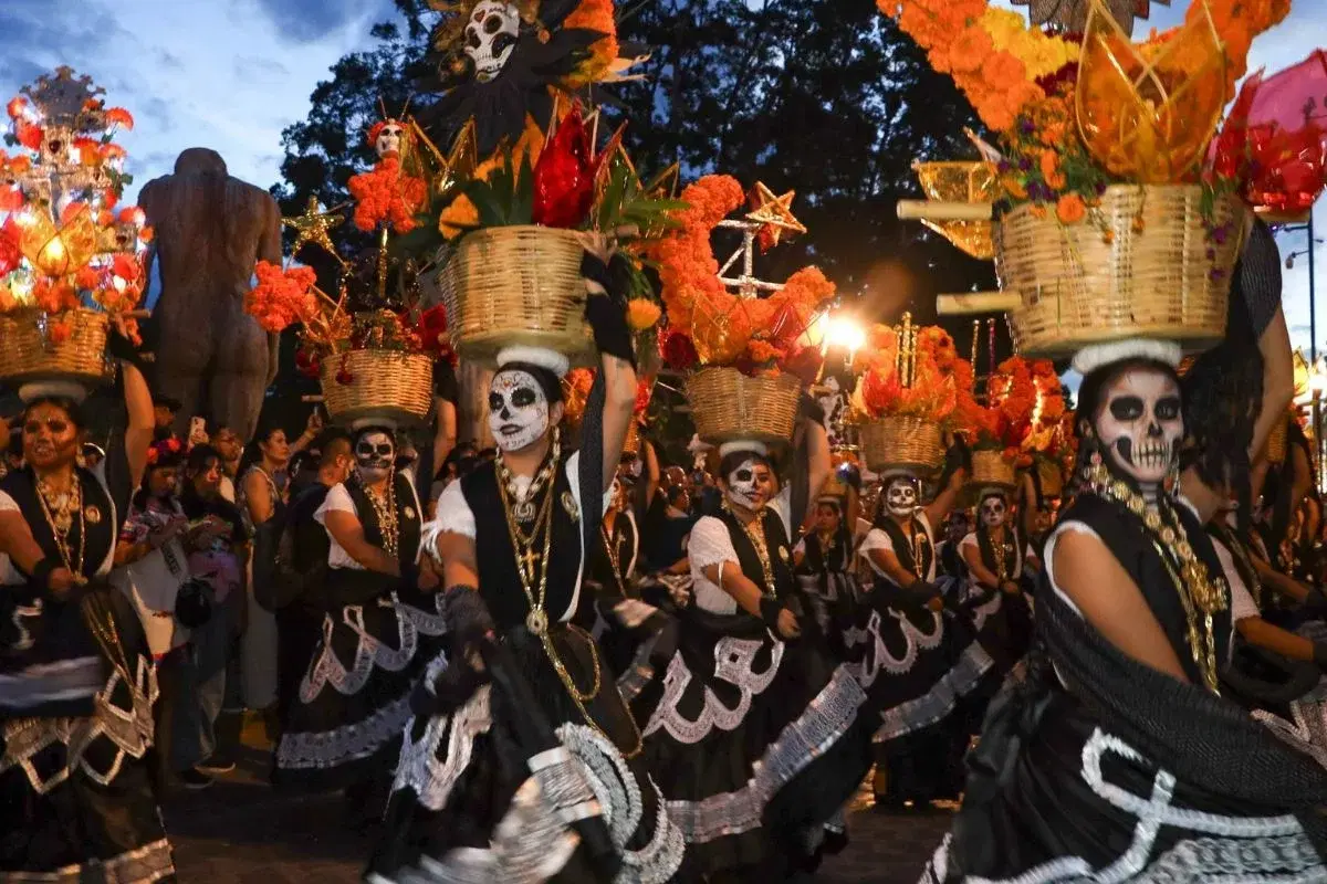 Comparsa de Día de Muertos en Oaxaca con mujeres catrinas que llevan canastas de cempasúchil sobre la cabeza